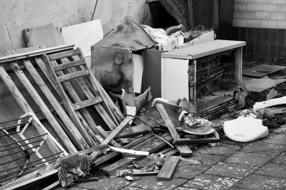 The image depicts a disorganized pile of discarded household items and debris placed on a paved outdoor area, likely part of a garden or driveway, adjacent to a brick wall. Prominently, there are two 